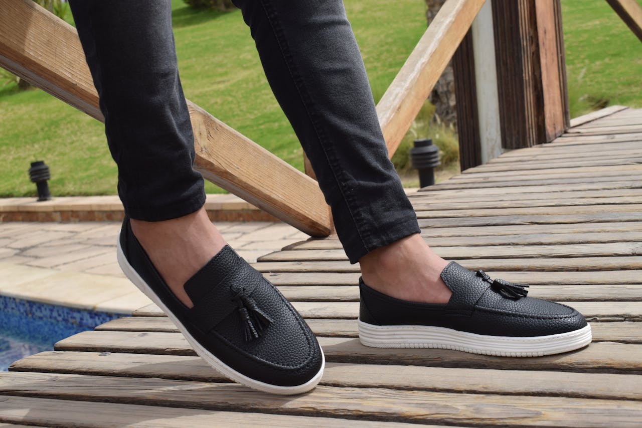 hero-img-02 Stylish black tassel loafers worn by a person standing on a wooden deck. Perfect for outdoor leisure fashion.
